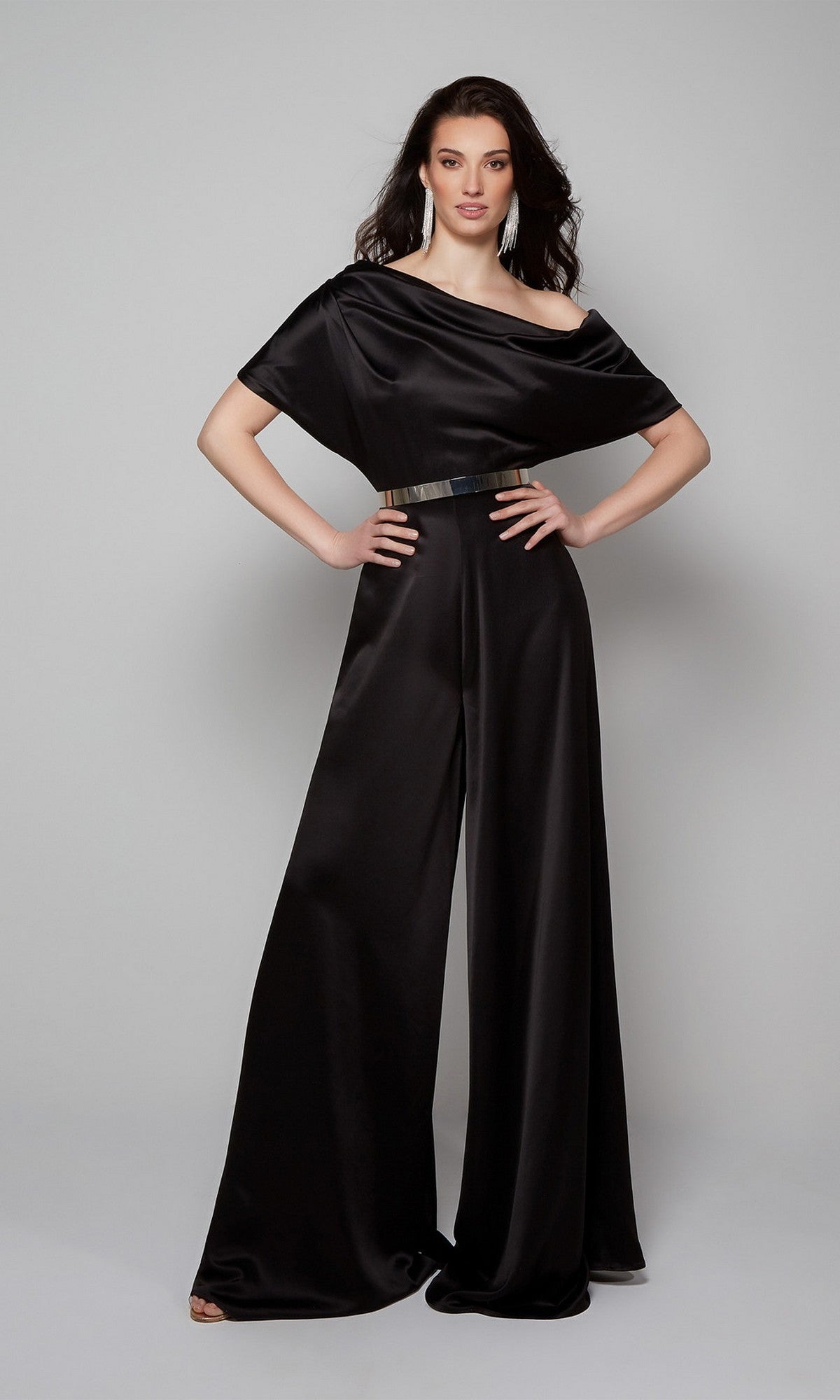 A woman poses in a studio against a plain background wearing the Long Formal Jumpsuit: Alyce 70019, a black wide-leg jumpsuit with short sleeves and a metallic belt, hands on hips.